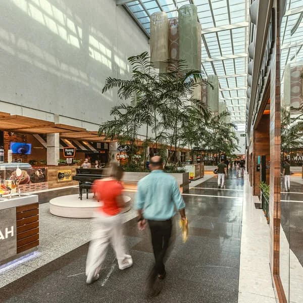 Corredor de um shopping Tacla, com pé-direito alto, plantas e vitrines